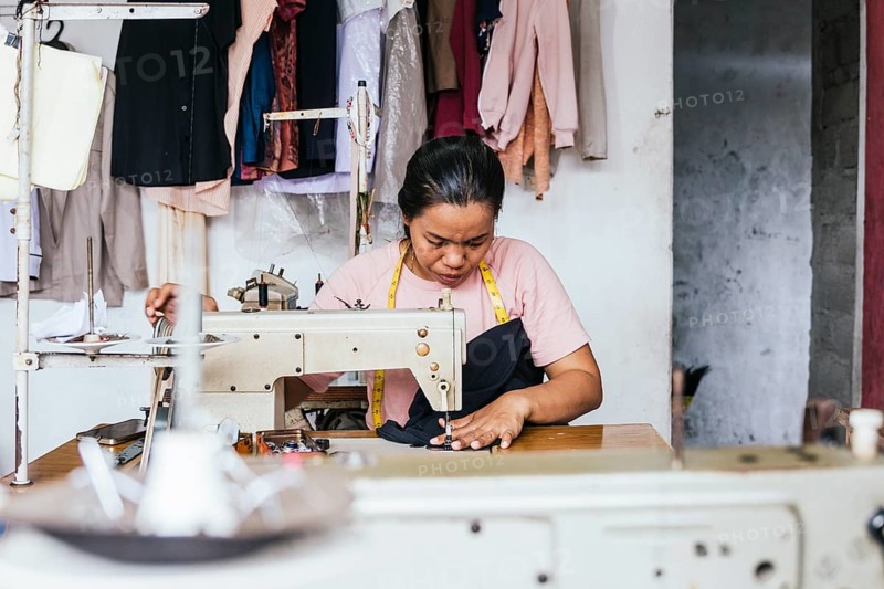 A asian seamstress is intently sewing in a machine, surrounded by hanging garments in a cozy tailor shop. Her workspace is organized with tools and clothing