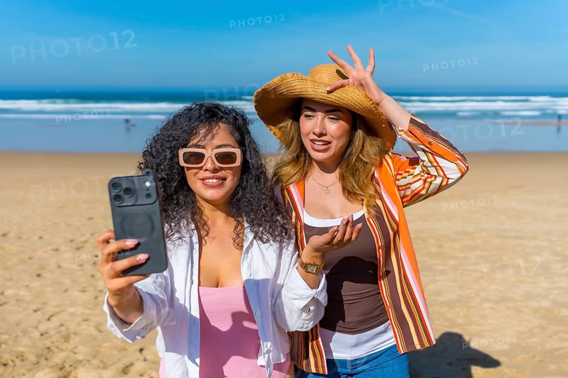 Two women friends smiling and posing for a selfie using a smartphone on a sandy beach with the ocean and blue sky in the background, enjoying their summer travel