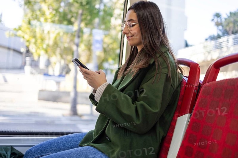 A serene woman wearing a green jacket sits in a red seat on public transport, engaged with her smartphone The setting reflects urban travel and modern connectivity