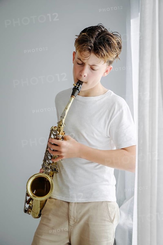 A young boy passionately playing the saxophone, wearing a white T shirt and beige pants, stands by a window with soft natural light streaming in, creating a peaceful atmosphere