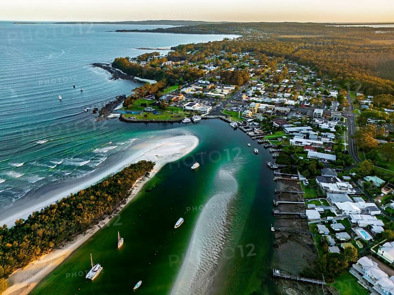 A stunning aerial view of a coastal town with a winding river, pristine beach, lush greenery, and residential buildings, capturing the serene beauty at sunset in Jervis Bay, Australia