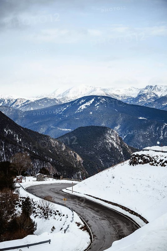 A winding road cuts through a snow covered valley amidst towering mountain peaks under a serene sky, offering a tranquil winter landscape that evokes peace in Andorra