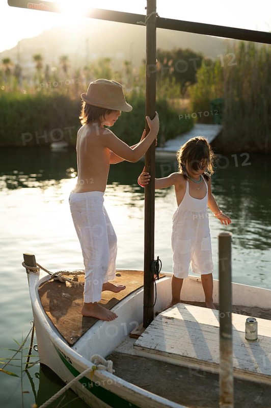 Siblings, a boy and a girl, play together on a boat in Andratx, Mallorca. They are both dressed in white, enjoying the serene surroundings of one of the Balearic Islands, Spain. The boy is looking away, holding a pole while the girl looks down