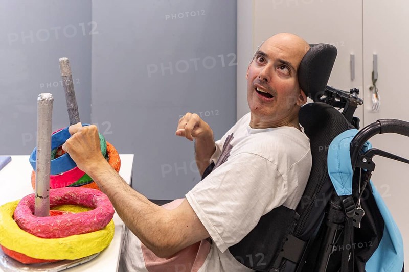 A man with a mental disability, seated in a wheelchair, enjoys a ring-stacking activity. His joyful expression and engagement highlight inclusion, creativity, and the importance of adaptive play