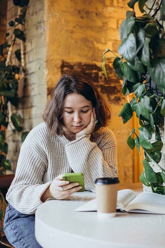 A young woman in a cozy sweater sits at a bookstore cafe table, scrolling on her smartphone An open book and a coffee cup are in front of her, surrounded by green plants