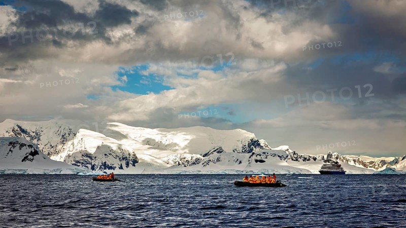 Two inflatable boats and a cruise ship in front of snowy mountains under a cloud cover at sea, people in an inflatable boat travelling on the sea in the Southern Ocean in Antarctica