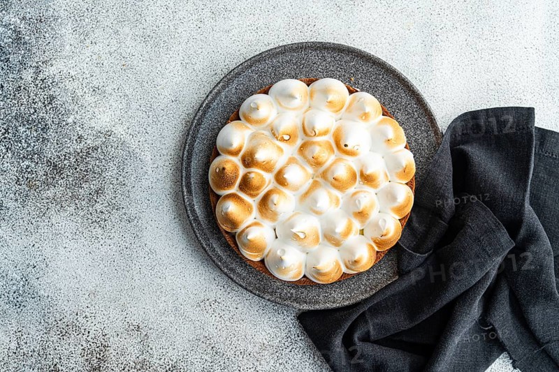 A lemon cake topped with toasted meringue sits on a dark plate, complemented by a cloth. The pie, with its golden peaks, offers a tempting visual on a textured background