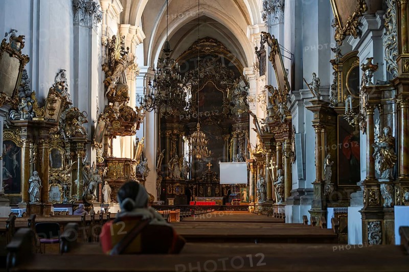 Grand interior of a baroque church featuring ornate golden altars and chandeliers with a person sitting in prayer in the foreground in Trzebnica, Poland