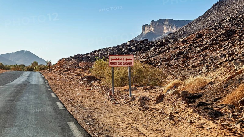 A tarmac country road runs through a dry desert landscape with a sign and mountains in the background, A drive through the mountainous landscape of the Sahara in Algeria