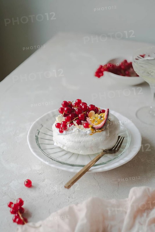 Beautifully arranged red currant and passionfruit pavlova on a white ceramic plate placed on a cloth-covered table. The dessert is garnished with fresh red currants and a sliced passionfruit, creating a visually appealing and appetizing image