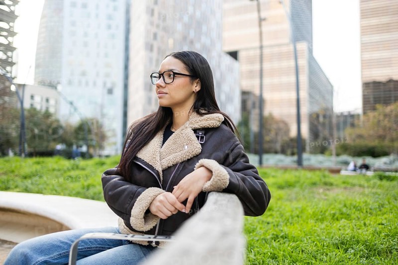 A woman in a cozy jacket sits on a bench, enjoying the urban scenery. She looks thoughtful amidst skyscrapers, embodying a balance of business and lifestyle