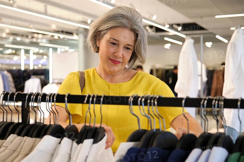Mature woman browsing through clothing on a rack in a modern store. Her gray hair and elegant yellow blouse add a touch of sophistication to the bright, well-lit environment