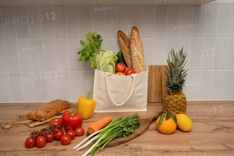 A reusable tote filled with fresh produce and baguettes sits on a wooden table. Surrounded by colorful fruits and vegetables, the setup promotes healthy, eco-conscious living