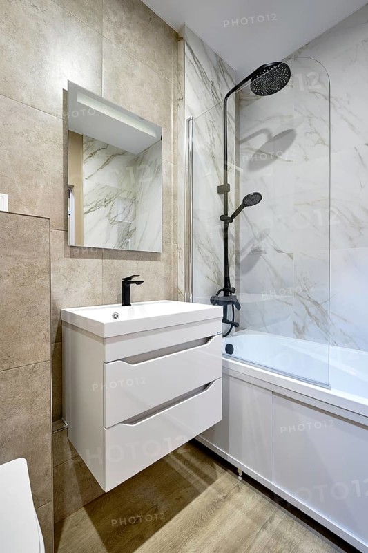 This modern bathroom features a sleek bathtub, a rainfall showerhead on a marble wall, and a minimalist vanity with drawers and a large mirror for both elegance and functionality