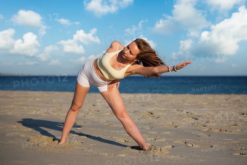 Adult woman practices Utthita Trikonasana, an extended triangle pose variation, on a sandy beach by the sea under a bright blue sky