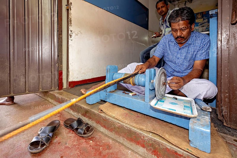 Man wearing a blue shirt operates a machine while sitting on the ground, The gemstone cutters in the city of Ratnapura in Sri Lanka