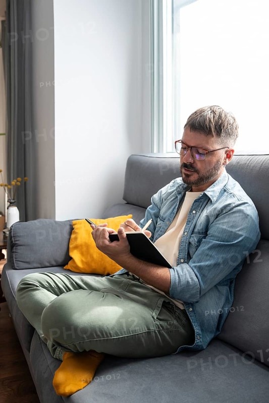 A man in casual attire sits cross-legged on a gray sofa, writing in a notebook. The living room is bright with natural light, featuring a yellow cushion and modern decor