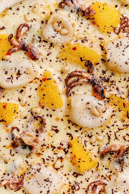 Close up view of a gourmet pizza topped with octopus, squid, slices of orange, mozzarella cheese, and scattered pine nuts, garnished with dark chocolate