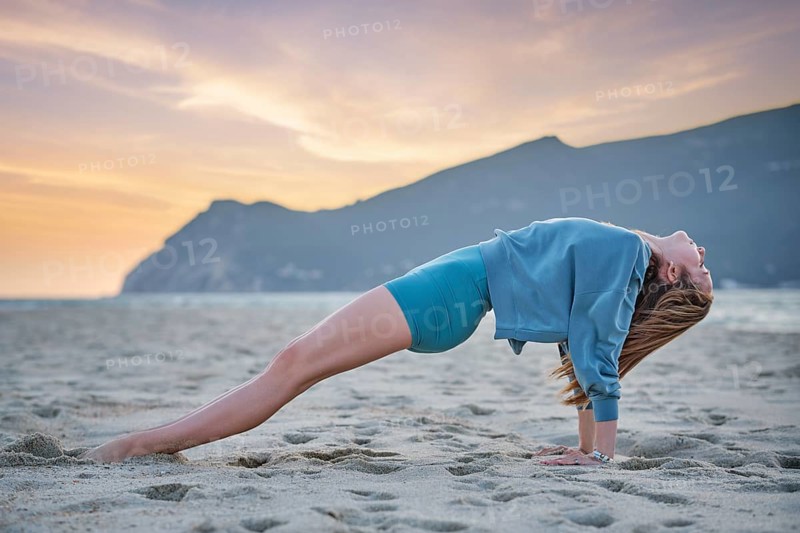A woman performs Purvottanasana, or Upward Plank Pose, on the sand at the beach. The sunset casts a warm glow, highlighting her strong core and serene expression amidst the natural beauty