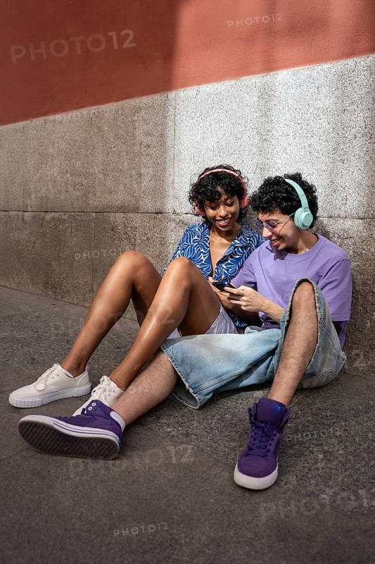 A joyful multiethnic LGBTQ+ couple sitting outdoors by a wall, listening to music and sharing earphones. They are smiling and relaxed, dressed in casual summer attire