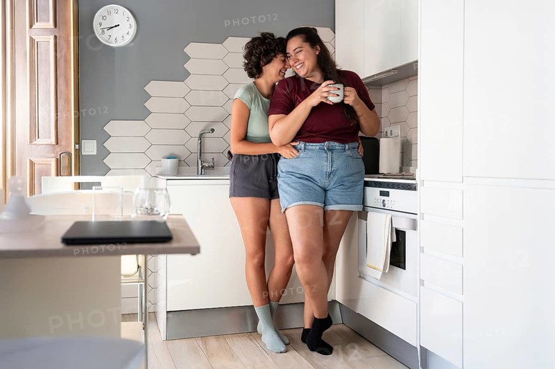 A happy lesbian couple shares a playful moment in a modern kitchen. One partner sits on the counter, feeding the other with a smile, showcasing a warm, intimate connection