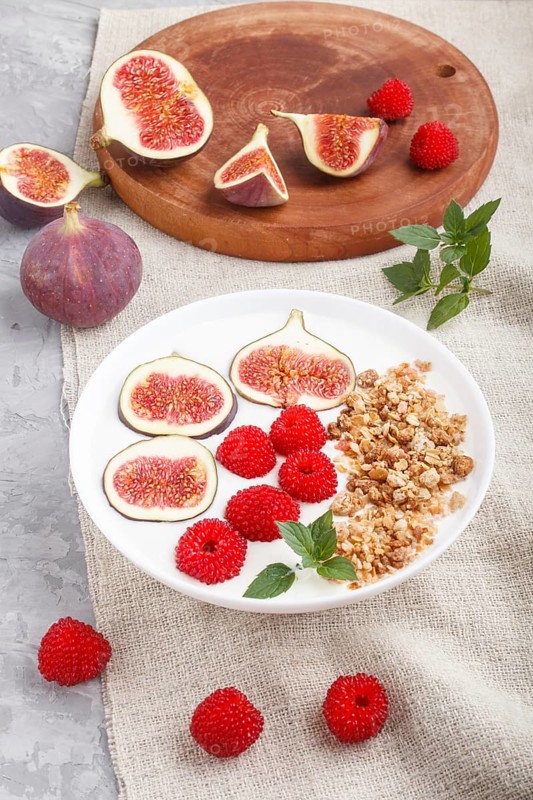 Yoghurt with raspberry, granola and figs in white plate on a gray concrete background and linen textile. side view, close up