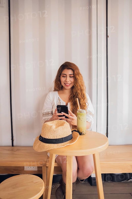 Young American Latin woman relaxing in a cafe, sipping a refreshing drink, and smiling while looking at her smartphone, exemplifying modern lifestyle and connectivity