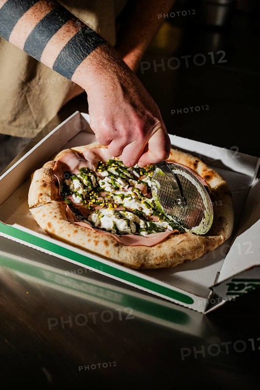 A close up shot of a hand cutting a freshly baked gourmet pizza in a takeout box using a metal pizza cutter. The pizza features rich toppings and a crispy crust