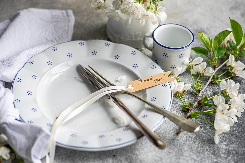 A beautifully set table with a white plate, cutlery, and a thank you note, adorned with delicate flowers and a cup. Ideal for special occasions or conveying gratitude