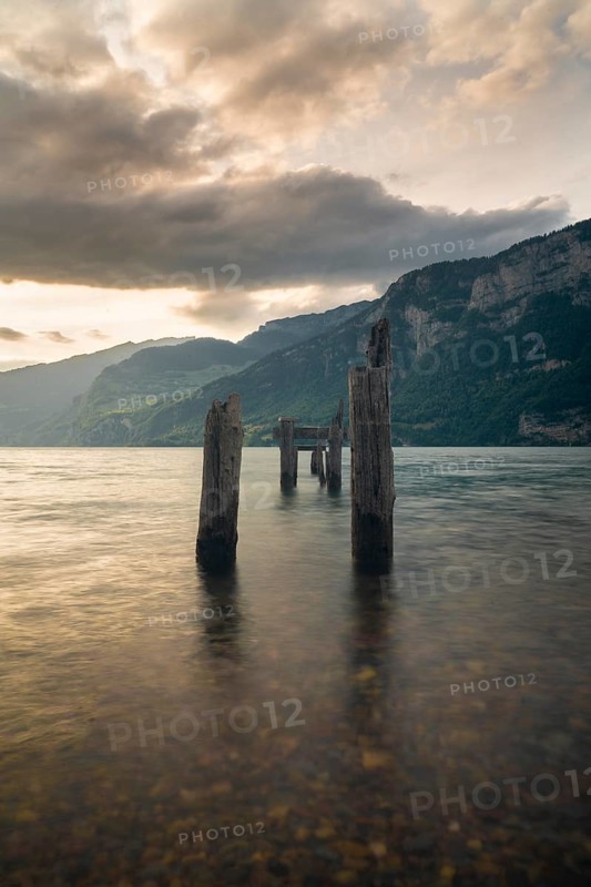 Old wooden pier posts rise from the still waters of Walensee in Switzerland, framed by dramatic mountains and a moody sunset sky. A peaceful yet powerful natural landscape