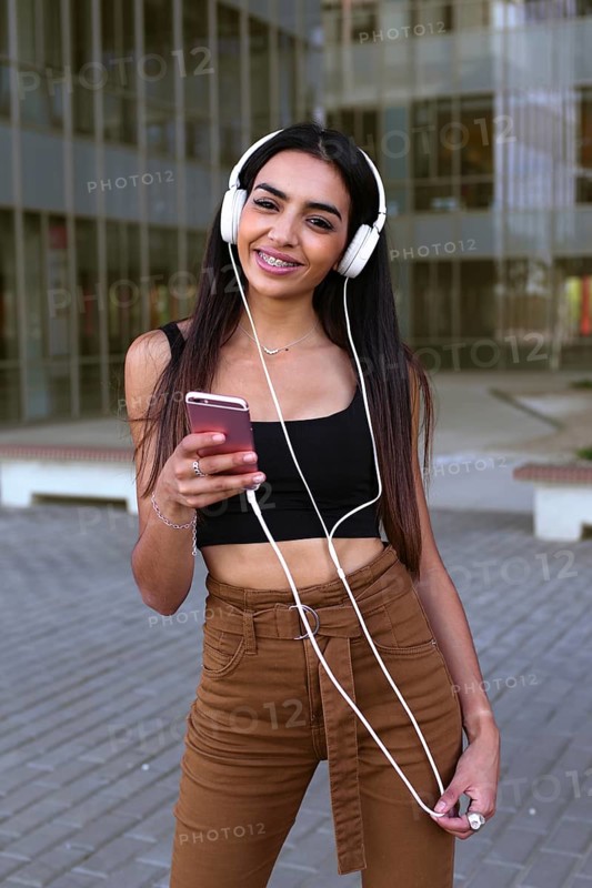 Young woman listening to music on headphones, holding her smartphone. She is outdoors, wearing casual clothing, embodying youthful energy