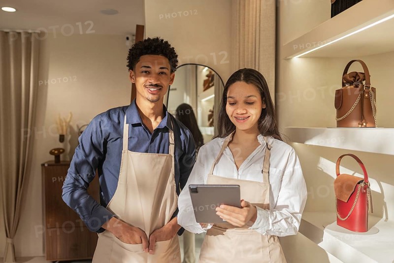 Two smiling store employees in aprons work together in a stylish, well lit bag store. They use a tablet to manage inventory and improve customer service. Handbags are displayed nearby