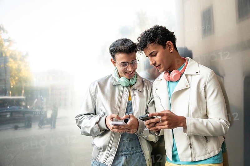 A young gay couple shares a joyful moment outdoors, smiling and interacting with their phones, exuding a sense of happiness and connection in a lively urban setting