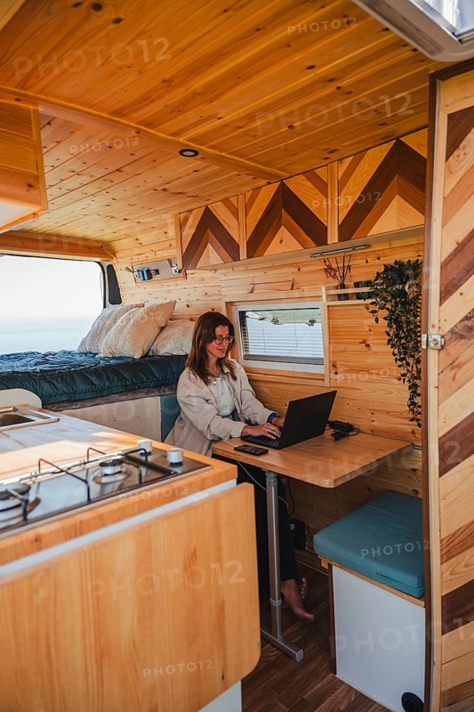 A woman works remotely on her laptop inside a cozy camper van, parked with a scenic view outside. The wood interior offers a warm and inviting workspace, ideal for travel