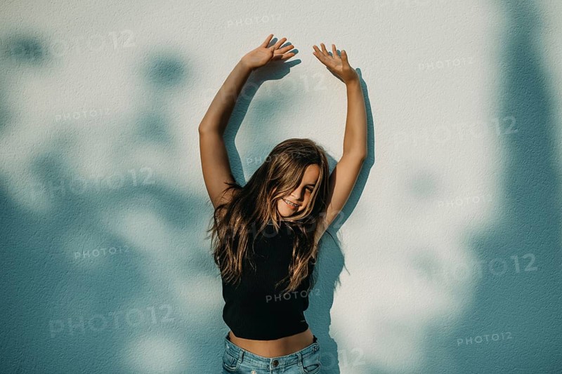 A teen girl stands with arms raised against a wall, with a joyful expression and shadows from tree branches casting patterns around her, creating a playful atmosphere