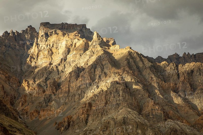 Stunning view of a rugged mountain range in Spiti Valley, India, under dramatic cloudy skies. The sunlight casts shadows, highlighting the rugged terrain and rocky textures