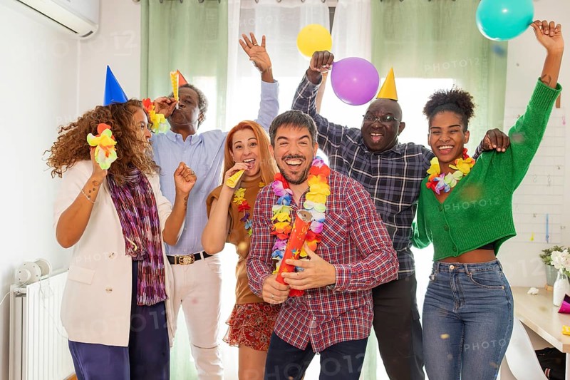 A vibrant group of friends celebrates a birthday indoors. They wear party hats and leis, hold balloons, and blow in noise makers, showcasing joy and togetherness