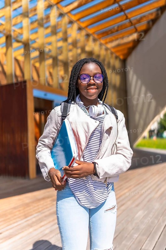 Vertical portrait of an African young woman standing outside the university smiling at camera holding folders