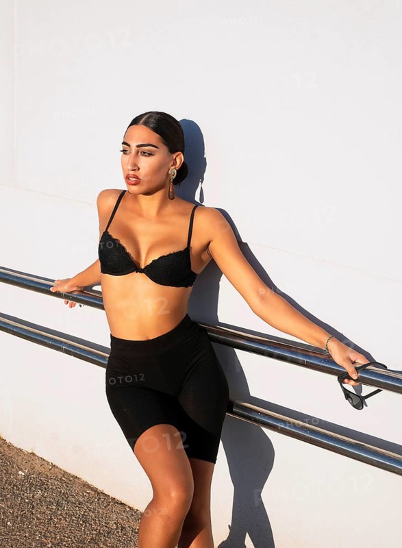 A confident woman leans against a railing, soaking in the bright sunlight. She wears black sportswear, exuding a sense of empowerment and style. Perfect for fashion and lifestyle themes