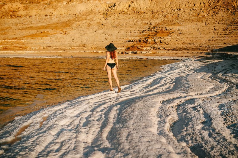 Back view of an unrecognizable woman in a bikini and wide-brimmed hat walking along the salt formations at the Dead Sea in Jordan