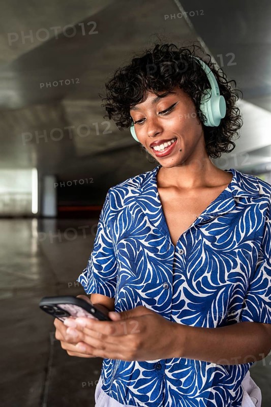 Woman with curly hair wearing blue patterned shirt listens to music on headphones while smiling and looking at her smartphone in a modern indoor setting