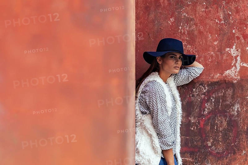 Stylish woman wearing a wide-brimmed hat and a fluffy vest leans against a textured, rustic wall. The setting exudes a blend of sophistication and urban charm