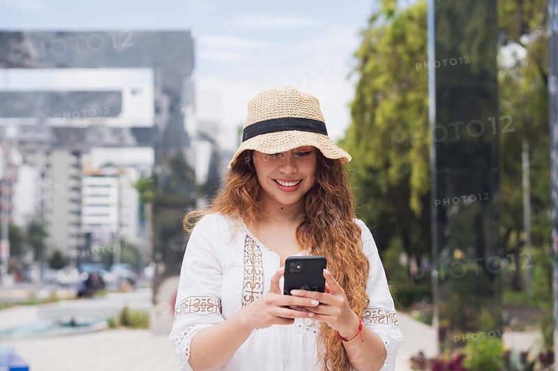 A young American Latin woman smiles while using her smartphone outdoors. Dressed in casual attire with a sun hat, she embodies a modern, relaxed lifestyle in an urban setting