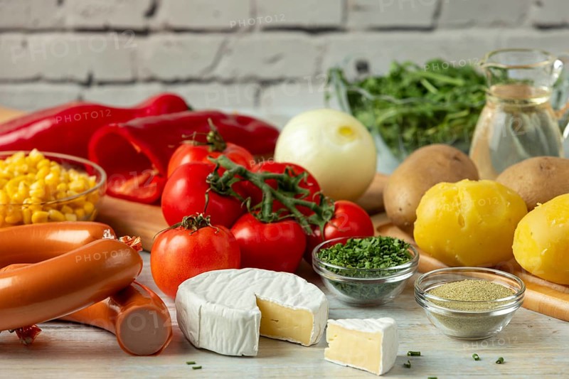Fresh ingredients such as potatoes, tomatoes, peppers, sausage, corn, onions, cheese and arugula in a bright environment