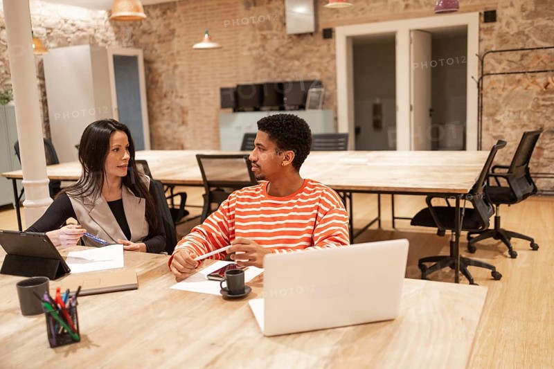 Two professionals collaborate in a modern coworking space, discussing ideas with laptops and documents Cozy atmosphere with exposed brick and contemporary furniture