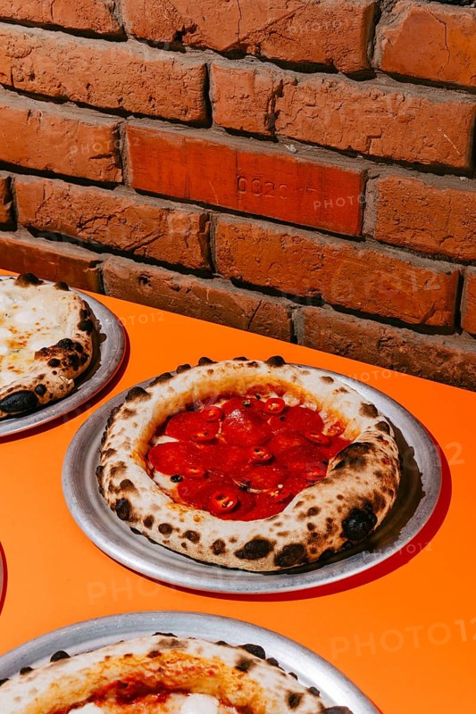 Wood-fired pepperoni pizza topped with salami, mozzarella, tomato sauce, and chili pepper. Served on a metal plate, featuring a golden crust and vibrant, spicy flavors