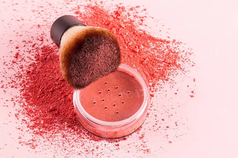 Close-up image of loose blush powder with a fluffy makeup brush on a pink background. The brush and powder create a soft and vibrant display ideal for beauty themes
