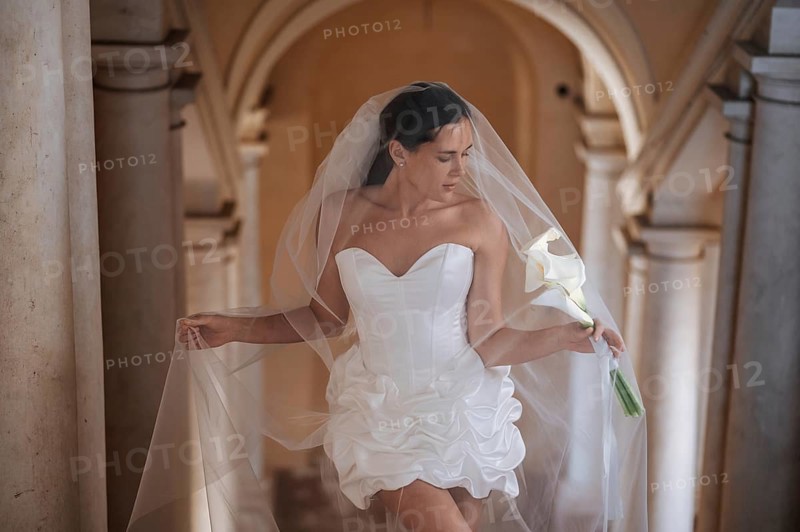 A bride in an elegant white dress and veil holds a calla lily in the historic setting of an ancient villa in Treia, Macerata, Italy. The scene exudes romance and tradition
