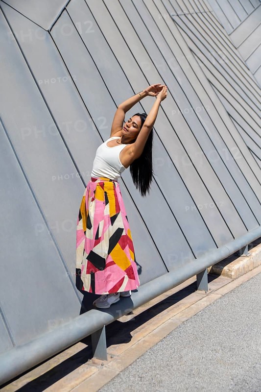 A woman in a vibrant, colorful skirt poses gracefully against a backdrop of striking modern architecture. Her outfit contrasts beautifully with the sleek urban setting