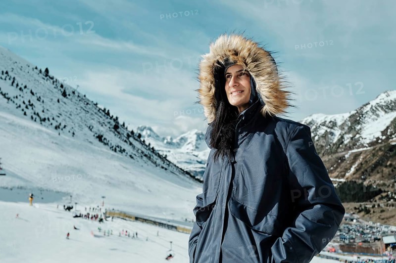 Woman in a fur-lined coat smiles against snowy mountains at Astun Ski Resort. Clear blue sky highlights the winter landscape of the Huesca Pyrenees, ideal for skiing